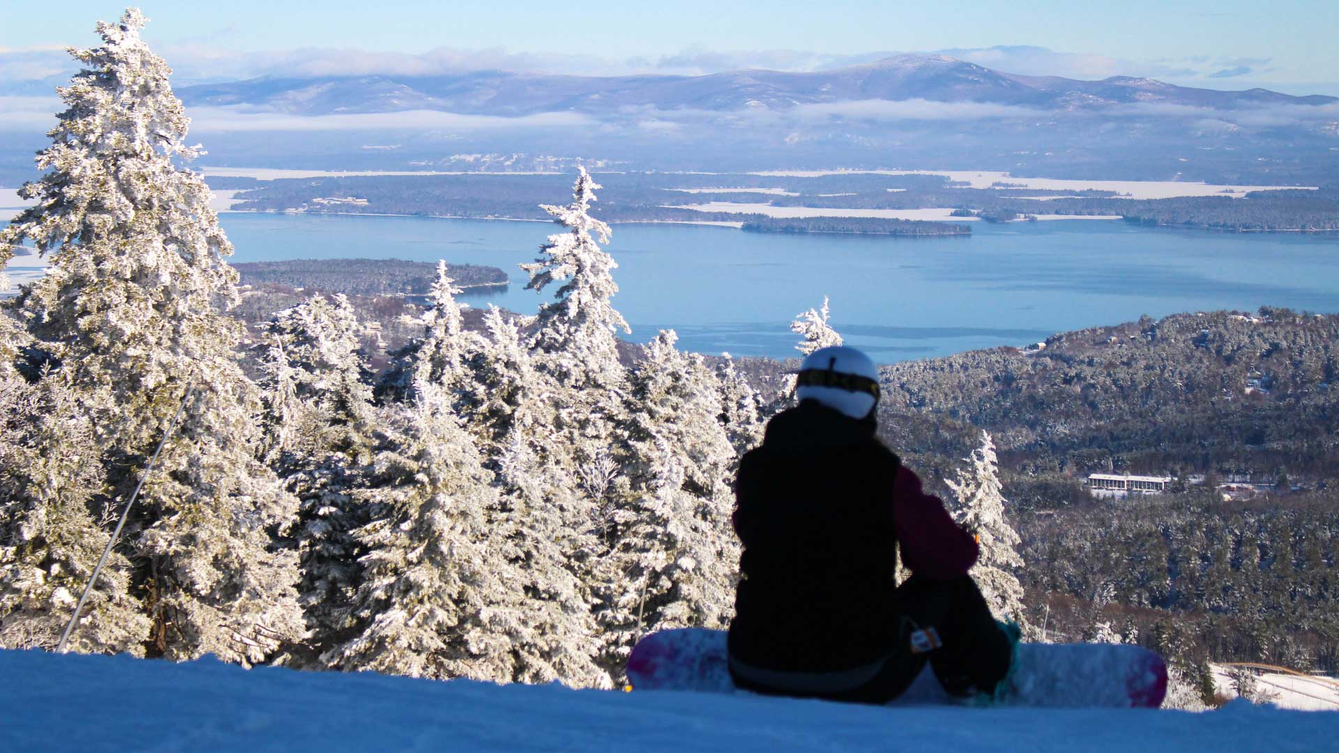 Snowboarder sits on a snowy mountainside overlooking valley, with lake and mountains in the distance