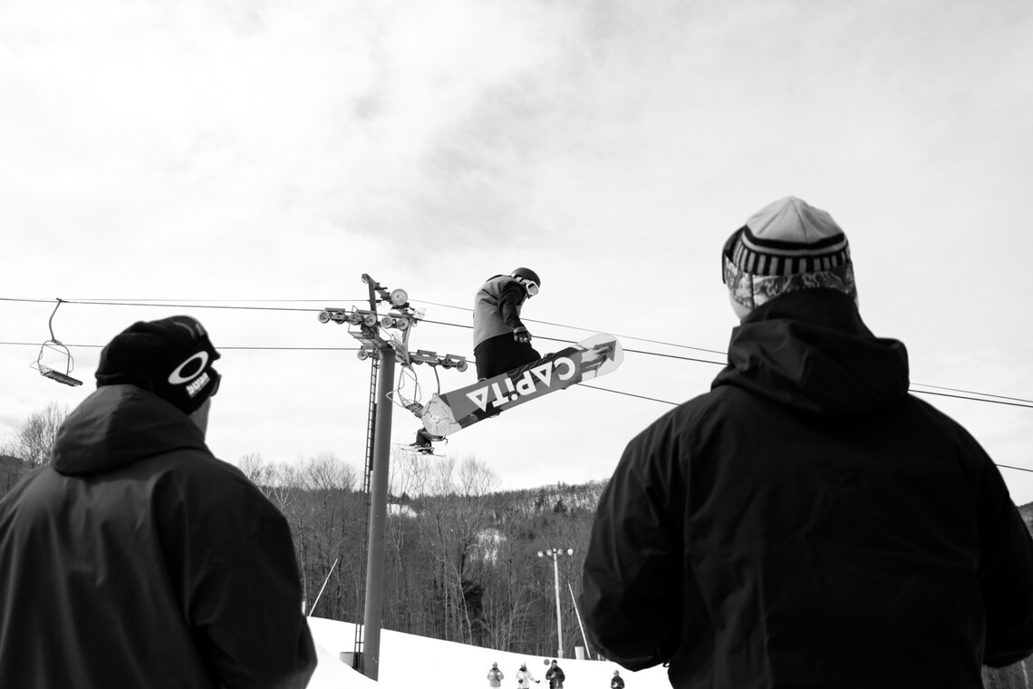Black and white photo of snowboarder jumping between two spectators.