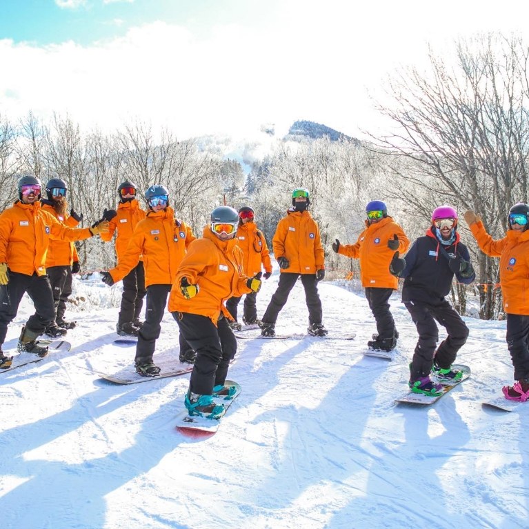 A group of snowboarders in orange jackets getting lessons